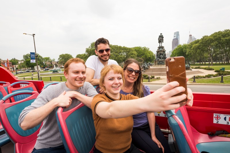 PST bus selfie A woman in a yellow shirt takes a selfie of herself and three friends on the top level of a City Sightseeing Philadelphia double decker tour bus.