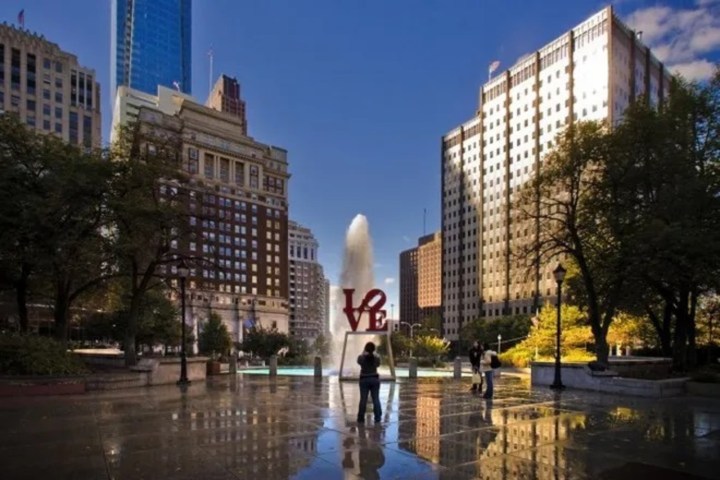 Urban park with LOVE sculpture, fountain, trees, and buildings under a blue sky.