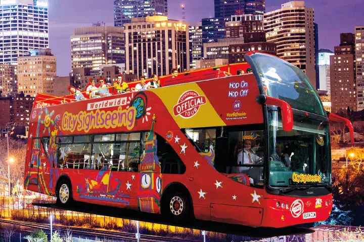 Red sightseeing bus with city skyline at dusk in the background.