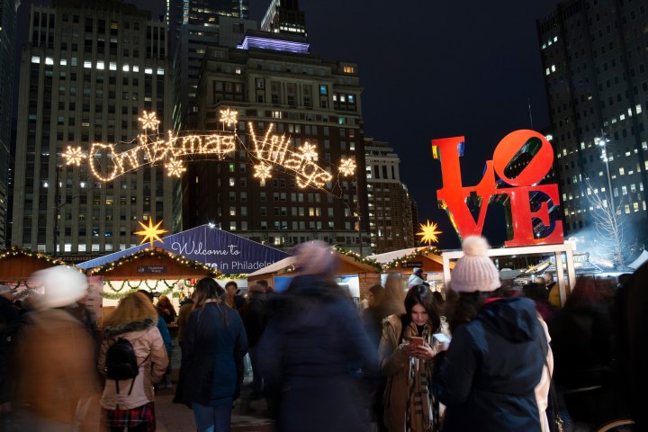 A crowd of people walking around the annual Christmas Village at Love Park. The Christmas Village sign made out of Christmas lights and the iconic LOVE sign are displayed above their heads.