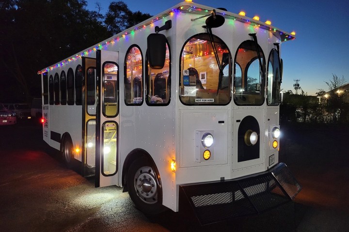 The exterior of a City Sightseeing Philadelphia trolley with the doors open wide. The roof is lined with multicolored Christmas lights that are all lit up in preparation for the Holiday Lights tour. Through the windows, we can see that the inside of the trolley is also lit up with Christmas lights.