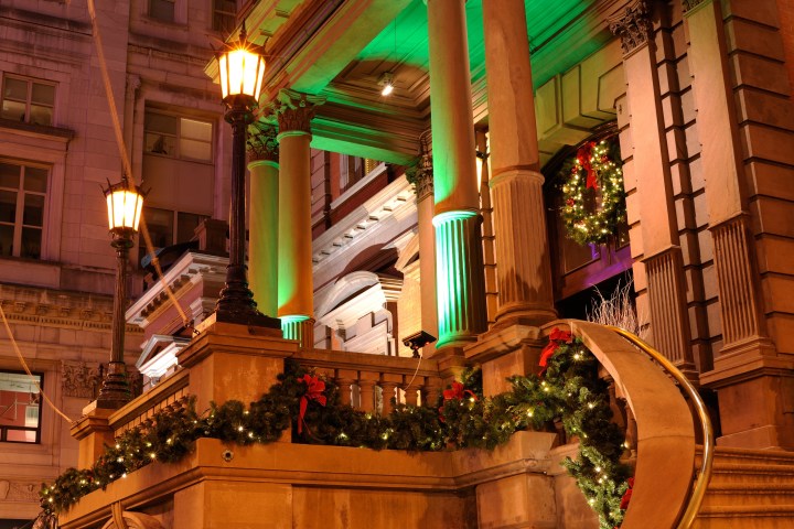 Philadelphia City Hall at night. The columns are lit up with green lights, and Christmas wreaths with white lights and red bows woven throughout are on the door and draped along the banister at the edge of the balcony and stairs.