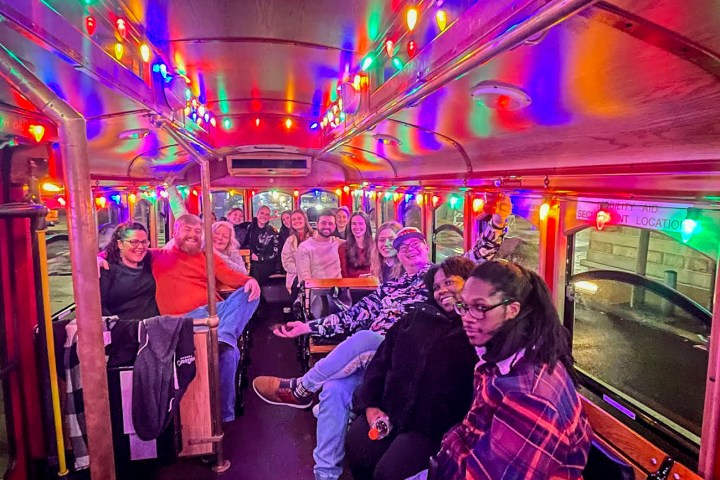 The interior of a City Sightseeing Philadelphia trolley full of happy, smiling people. The ceiling and windows are lined with multicolored Christmas lights that are all lit up in preparation for the Holiday Lights tour.