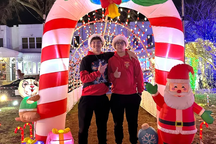 Two people in Santa hats under a candy cane arch with colorful lights and inflatable Santa, gifts.