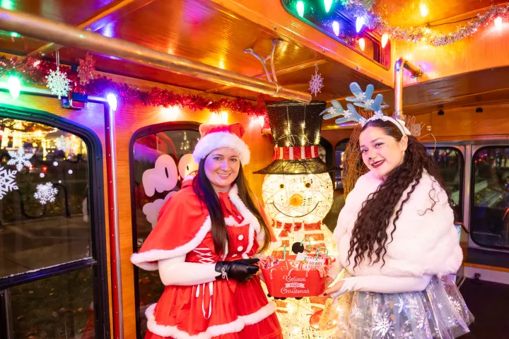 Two women in festive outfits pose inside a decorated trolley with a snowman light display.