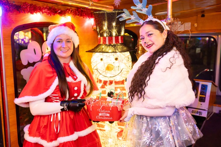Two women in festive costumes posing with a lit snowman decoration holding a Christmas-themed box.