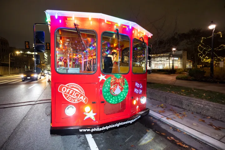 Red tour trolley decorated with colorful lights and a wreath, parked on a city street at night.