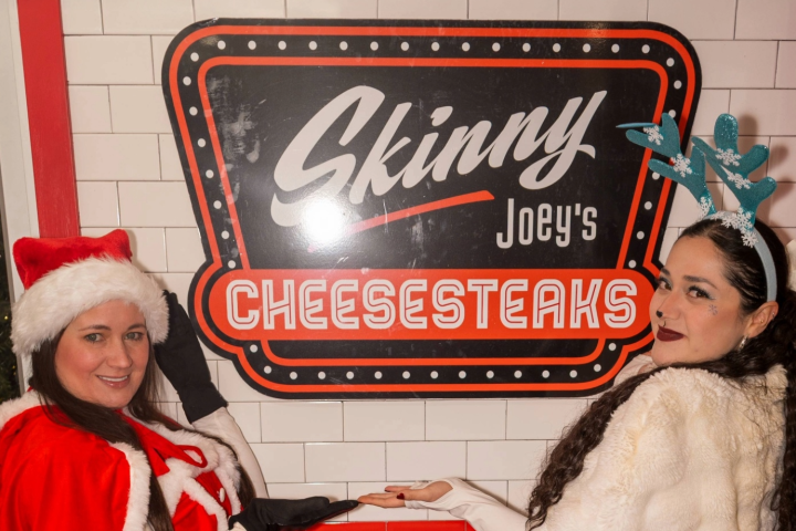 Two women in festive outfits pose by a sign for 'Skinny Joey's Cheesesteaks.'