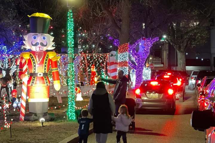 People walk near festive lights and a large nutcracker statue at night with cars nearby.